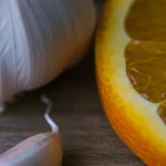 Primo piano macro di uno spicchio d'aglio fresco e una fetta d'arancia vibrante affiancati su una superficie di legno rustico, illuminazione controllata e morbida, alta definizione, lente macro 90mm, a simboleggiare l'unione protettiva di allicina ed esperidina per la fertilità maschile contro le tossine ambientali.