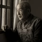Ritratto fotografico di un uomo anziano cinese, circa 70 anni, seduto vicino a una finestra con luce soffusa, sguardo pensieroso ma sereno; prime lens 35mm, profondità di campo, tonalità duotone seppia e grigio.