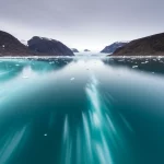 Fotografia paesaggistica di un fiordo artico (Kongsfjorden, Svalbard), obiettivo grandangolare 15mm, lunga esposizione per acqua liscia, messa a fuoco nitida, con un ghiacciaio marino che si ritira sullo sfondo e pennacchi di sedimento visibili nell'acqua turchese sotto un cielo artico luminoso ma nuvoloso.