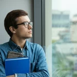 Primo piano fotografico di uno studente universitario, forse proveniente da un percorso VET, che guarda fuori da una finestra del campus con espressione pensierosa e leggermente stanca, simboleggiando il mix di stress accademico e speranza. Obiettivo 50mm, luce naturale morbida, profondità di campo media che sfoca leggermente lo sfondo del campus.