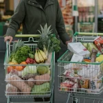 Una persona adulta indecisa davanti a due carrelli della spesa: uno pieno di frutta, verdura e cibi freschi, l'altro pieno di snack confezionati, bibite gassate e cibi pronti ultra-processati. Prime lens, 35mm portrait, depth of field, luce naturale morbida che illumina la scena.