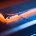 Concetto astratto del trattamento termico di una lega metallica, rappresentato da un bagliore caldo (arancione) che sfuma verso un blu freddo su una superficie metallica lucida, fotografia con obiettivo prime 35mm, effetto duotone blu e arancione, profondità di campo che crea un bokeh suggestivo.