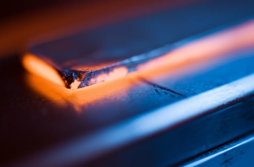 Concetto astratto del trattamento termico di una lega metallica, rappresentato da un bagliore caldo (arancione) che sfuma verso un blu freddo su una superficie metallica lucida, fotografia con obiettivo prime 35mm, effetto duotone blu e arancione, profondità di campo che crea un bokeh suggestivo.