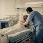 Fotografia realistica in stile reportage: un paziente con lesione midollare a letto in una stanza d'ospedale, assistito da un infermiere attento durante una procedura vicino al letto. Luce soffusa dalla finestra, obiettivo 35mm, profondità di campo che sfoca leggermente lo sfondo per concentrarsi sull'interazione. Toni caldi e freddi duotone (blu e ocra).