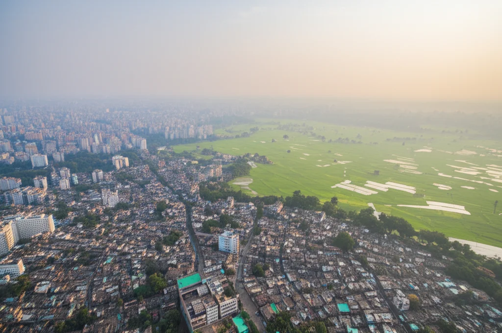 Veduta aerea grandangolare del Bangladesh (obiettivo 18mm, landscape wide angle) che mostra un mix di aree urbane dense e campagne verdi sotto un cielo con una leggera foschia atmosferica visibile, rappresentante l'AOD. La luce solare filtra debolmente attraverso la foschia, creando un'atmosfera suggestiva ma che allude all'inquinamento. Messa a fuoco nitida su tutto il paesaggio, lunga esposizione per eventuali nuvole soffici.