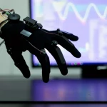Immagine fotorealistica di una mano umana che indossa un data glove high-tech nero, con fili sottili e sensori visibili. La mano è sospesa sopra una superficie liscia e scura, e le dita sono leggermente flesse come se stessero interagendo con un'interfaccia invisibile. Sullo sfondo, uno schermo di computer mostra un cursore astratto e linee di dati, leggermente sfocato per mantenere il focus sulla mano. L'illuminazione è drammatica, quasi da studio, con un obiettivo prime 35mm che crea una leggera profondità di campo, enfatizzando la connessione tra l'azione umana e la tecnologia.