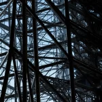 Visualizzazione fotorealistica di una complessa struttura reticolare metallica, simile a una torre o ponte, illuminata da luce drammatica che ne evidenzia la geometria ottimizzata. Obiettivo grandangolare 20mm, messa a fuoco nitida, dettagli elevati della struttura.