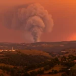 Fotografia paesaggistica grandangolare, 10mm, che cattura un vasto paesaggio collinare del nord del Portogallo al tramonto, con una colonna di fumo di un incendio boschivo che si alza in lontananza sotto un cielo infuocato, lunga esposizione per rendere il fumo etereo, messa a fuoco nitida sull'orizzonte.