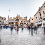 Una folla stilizzata di turisti in una piazza iconica italiana come Piazza San Marco a Venezia, alcuni disegnati in modo solido e altri come sagome trasparenti o 'fantasma' per rappresentare il turismo non osservato e sommerso. Obiettivo grandangolare 20mm per catturare l'ampiezza della piazza, messa a fuoco nitida sull'intera scena, alcuni turisti in movimento leggermente sfocato per dare dinamicità.