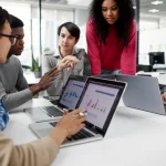 Gruppo diversificato di giovani futuri insegnanti di matematica (uomini e donne, età 20-25 anni) partecipano attivamente a un workshop sull'intelligenza artificiale in un'aula universitaria moderna e luminosa. Sono riuniti attorno a un tavolo, interagendo con laptop e tablet che mostrano interfacce AI complesse e grafici matematici. Alcuni discutono tra loro, altri sono concentrati sugli schermi. L'atmosfera è di apprendimento collaborativo e riflessione critica. Obiettivo zoom 24-70mm per catturare la dinamica del gruppo, luce ambientale morbida e naturale.