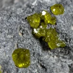 Immagine fotorealistica: Scatto macro ravvicinato, obiettivo 100mm, di diversi minuscoli cristalli di olivina giallo-verdastri incastonati in un frammento di suolo lunare grigio scuro, high detail che rivela la struttura cristallina, precise focusing, illuminazione controllata tipo studio che enfatizza la texture e le variazioni di colore.