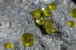 Immagine fotorealistica: Scatto macro ravvicinato, obiettivo 100mm, di diversi minuscoli cristalli di olivina giallo-verdastri incastonati in un frammento di suolo lunare grigio scuro, high detail che rivela la struttura cristallina, precise focusing, illuminazione controllata tipo studio che enfatizza la texture e le variazioni di colore.