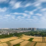 Veduta aerea grandangolare (10mm) di un paesaggio cinese che mostra la transizione tra una zona urbana densa e una rurale agricola sotto un cielo parzialmente nuvoloso, simboleggiando i cambiamenti climatici e l'impatto umano, long exposure per nuvole fluide.