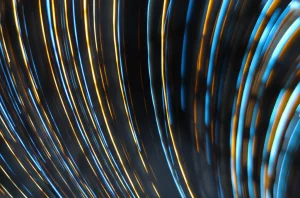 Visualizzazione astratta di traiettorie luminose curve nello spaziotempo, che interagiscono con superfici geometriche complesse rappresentanti condizioni al contorno non standard. Effetti di lente gravitazionale suggeriti. Obiettivo grandangolare, 15mm, lunga esposizione per scie luminose, messa a fuoco nitida sulle superfici.