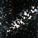 Immagine fotorealistica di un reattore a letto impaccato trasparente riempito con particelle di biochar nero di varie dimensioni e forme irregolari, con un fascio di luce che ne illumina la struttura interna complessa e gli spazi vuoti, obiettivo macro 80mm, illuminazione drammatica laterale, alta definizione, profondità di campo ridotta.