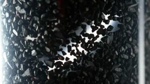 Immagine fotorealistica di un reattore a letto impaccato trasparente riempito con particelle di biochar nero di varie dimensioni e forme irregolari, con un fascio di luce che ne illumina la struttura interna complessa e gli spazi vuoti, obiettivo macro 80mm, illuminazione drammatica laterale, alta definizione, profondità di campo ridotta.