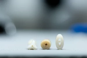 Immagine macro ad alta definizione di tre diversi connettori per protesi dentali (triangolare, quadrato, ovale) realizzati in Zirconia bianca, PEEK beige e FRC traslucido, illuminazione da studio dentistico, obiettivo macro 90mm, focus preciso sulle geometrie dei connettori, sfondo sfocato di laboratorio odontotecnico.
