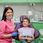 Una dentista pediatrica indossa un camice colorato (rosa) e sorride a un bambino seduto sulla poltrona dentistica in uno studio moderno e accogliente. Fotografia stile ritratto, obiettivo 35mm, luce morbida, profondità di campo che sfoca leggermente lo sfondo, colori vivaci ma naturali.