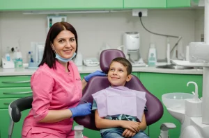 Una dentista pediatrica indossa un camice colorato (rosa) e sorride a un bambino seduto sulla poltrona dentistica in uno studio moderno e accogliente. Fotografia stile ritratto, obiettivo 35mm, luce morbida, profondità di campo che sfoca leggermente lo sfondo, colori vivaci ma naturali.
