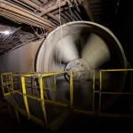 Fotografia wide-angle 15mm di un imponente ventilatore assiale industriale in funzione all'interno di una galleria mineraria scarsamente illuminata, polvere sospesa nell'aria illuminata da un fascio di luce, messa a fuoco nitida sul ventilatore, lunga esposizione per mostrare il movimento sfocato delle pale.