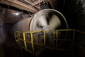 Fotografia wide-angle 15mm di un imponente ventilatore assiale industriale in funzione all'interno di una galleria mineraria scarsamente illuminata, polvere sospesa nell'aria illuminata da un fascio di luce, messa a fuoco nitida sul ventilatore, lunga esposizione per mostrare il movimento sfocato delle pale.