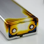 Immagine fotorealistica: Batteria agli ioni di litio parzialmente smontata su un banco di lavoro, che rivela chiaramente l'involucro in lega di alluminio 5052. Un angolo dell'involucro mostra sottili segni di stress termico o leggera carbonizzazione, alludendo visivamente al rischio di combustione. Obiettivo macro, 70mm, illuminazione controllata che enfatizza le texture metalliche e i dettagli, alta definizione.