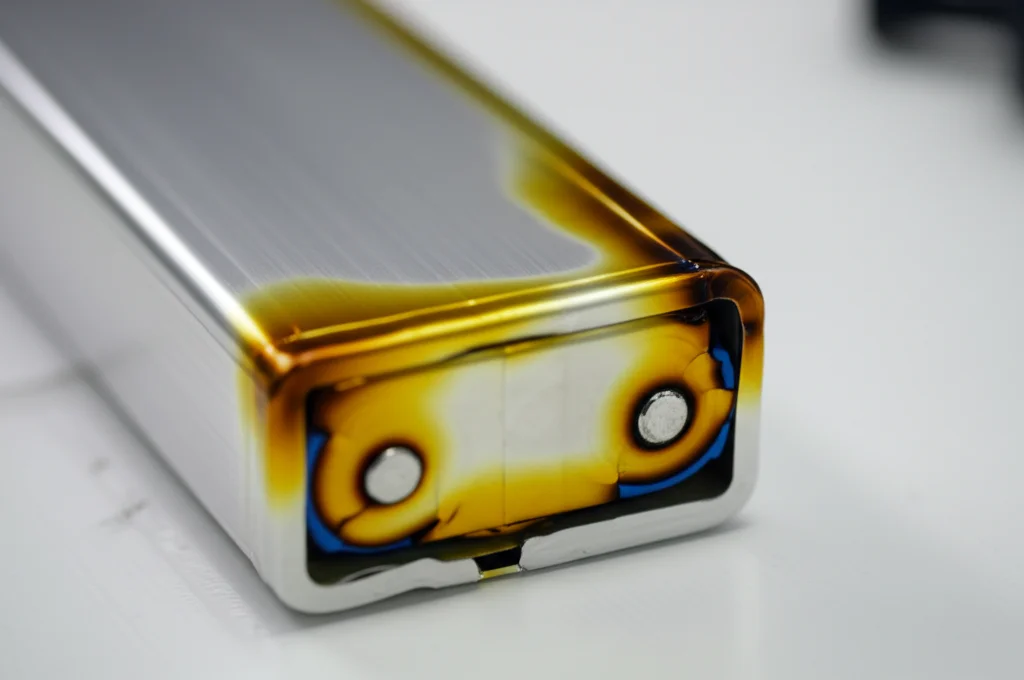 Immagine fotorealistica: Batteria agli ioni di litio parzialmente smontata su un banco di lavoro, che rivela chiaramente l'involucro in lega di alluminio 5052. Un angolo dell'involucro mostra sottili segni di stress termico o leggera carbonizzazione, alludendo visivamente al rischio di combustione. Obiettivo macro, 70mm, illuminazione controllata che enfatizza le texture metalliche e i dettagli, alta definizione.
