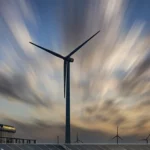 Immagine fotorealistica di una moderna centrale elettrica a ciclo combinato affiancata da turbine eoliche e pannelli solari al tramonto, vista grandangolare 18mm, lunga esposizione per un cielo drammatico con nuvole striate, simboleggia la pianificazione strategica per la sostenibilità energetica bilanciando diverse tecnologie.