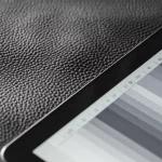 Immagine macro ad alto dettaglio di un campione di pelle pregiata sottoposto a test di sfregamento, con accanto una scala dei grigi digitale visualizzata su un tablet moderno. Illuminazione controllata da laboratorio che enfatizza la texture della pelle e la precisione della scala digitale. Obiettivo macro 90mm, messa a fuoco precisa.