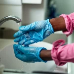 Fotografia realistica di un operatore sanitario in Camerun che indossa guanti monouso blu e si lava accuratamente le mani in un lavandino d'ospedale. Luce controllata, obiettivo macro 60mm, alta definizione, messa a fuoco precisa sui dettagli delle gocce d'acqua e della schiuma di sapone.