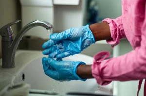 Fotografia realistica di un operatore sanitario in Camerun che indossa guanti monouso blu e si lava accuratamente le mani in un lavandino d'ospedale. Luce controllata, obiettivo macro 60mm, alta definizione, messa a fuoco precisa sui dettagli delle gocce d'acqua e della schiuma di sapone.