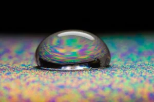 Macro fotografia, obiettivo 100mm, di una singola goccia di collirio trasparente che si trasforma in un gel viscoso mentre tocca una superficie iridescente che simboleggia la superficie oculare, rappresentando un sistema avanzato di rilascio oftalmico di CoQ10. Alta definizione, illuminazione da studio controllata, messa a fuoco precisa sulla transizione liquido-gel.