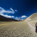 Fotografia paesaggistica, grandangolo 12mm, che cattura l'immensità delle montagne tibetane nella regione di Shannan sotto un cielo azzurro intenso, con una donna in abiti tradizionali che cammina su un sentiero in lontananza. Luce solare diretta, messa a fuoco nitida su tutto il panorama, lunga esposizione per rendere le nuvole soffici.
