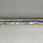 Primo piano di una saldatura perfetta e lucida tra una lastra di lega di magnesio grigiastra opaca e una lastra di acciaio inossidabile brillante con un sottile strato intermedio di nichel visibile. La saldatura è stata realizzata con tecnologia laser-ultrasuoni. Dettagli macro con obiettivo 100mm, illuminazione controllata da laboratorio che esalta le texture dei metalli, alta definizione, messa a fuoco precisa sulla linea di giunzione.