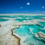 Fotografia di paesaggio, obiettivo grandangolare 12mm, lunga esposizione per rendere l'acqua setosa, che mostra una moderna piattaforma carbonatica tropicale con secche oolitiche e piane di marea esposte sotto un cielo azzurro intenso, evocando l'ambiente deposizionale della Formazione Longwangmiao nel Cambriano.