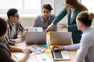 Gruppo diversificato di studenti universitari che collaborano attorno a un tavolo, alcuni usano laptop con grafici AI, altri discutono animatamente, luce naturale da finestra, obiettivo zoom 24-70mm, scena dinamica.