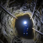 Immagine fotorealistica dell'interno di un cantiere di scavo profondo nella miniera d'oro di Xiadian. Si vedono le pareti rocciose grezze, umide e scintillanti sotto la luce delle lampade. Un minatore in tuta da lavoro, con casco e lampada frontale accesa, è visibile in secondo piano, intento a lavorare con attrezzature minerarie. L'aria sembra densa e pesante. Obiettivo zoom 24-70mm impostato a 28mm per una visione ampia ma immersiva, profondità di campo media, illuminazione mista tra le lampade frontali e luci di lavoro fisse che creano ombre profonde, evidenziando l'ambiente difficile.
