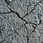Primo piano fotorealistico di un campione di CPB (Cemented Paste Backfill) fratturato dopo un test di compressione. Si vedono chiaramente le particelle aggregate (ganga di carbone), la matrice cementizia e le micro-fratture interne che attraversano il materiale. Obiettivo macro 100mm, alta definizione, illuminazione da studio controllata per enfatizzare la texture e il degrado strutturale, profondità di campo ridotta per isolare il dettaglio della frattura.