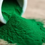 Primo piano fotorealistico, obiettivo macro 100mm, di vibrante polvere di Spirulina platensis verde scuro che si riversa da un piccolo mortaio di ceramica bianca su una superficie di legno rustico. Luce laterale morbida che ne esalta la texture finissima e il colore intenso. Alta definizione, messa a fuoco precisa sulla polvere, sfondo leggermente sfocato.