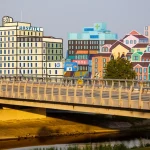 Fotografia realistica di un ponte moderno che collega due sponde diverse, una rappresentante un ospedale stilizzato e l'altra una comunità cittadina vivace, obiettivo grandangolare 24mm, luce calda del tardo pomeriggio, a simboleggiare il collegamento e la partnership cruciale tra sanità e territorio per la cura delle dipendenze.
