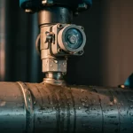 Primo piano di un sensore di vibrazione high-tech montato su una tubatura d'acqua industriale metallica, con goccioline d'acqua visibili sulla superficie del tubo vicino al sensore, obiettivo prime 35mm, profondità di campo ridotta per focalizzare sul sensore e le gocce, illuminazione laterale che crea ombre definite.