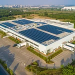 Veduta aerea con obiettivo grandangolare 15mm di una moderna fabbrica indiana eco-sostenibile perfettamente integrata in un paesaggio verde lussureggiante, con pannelli solari sui tetti e camion elettrici per la logistica visibili nel piazzale, focus nitido su tutta la scena, luce naturale del tardo pomeriggio che crea lunghe ombre.