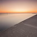 Fotografia grandangolare, 20mm, di un'alta marea primaverile che raggiunge il limite di una passeggiata lungomare in cemento al tramonto. L'acqua è calma ma molto alta, quasi a livello della passeggiata. Il cielo riflette colori caldi sull'acqua. Focus nitido sull'interfaccia acqua-cemento, lunga esposizione per acqua setosa, atmosfera suggestiva.