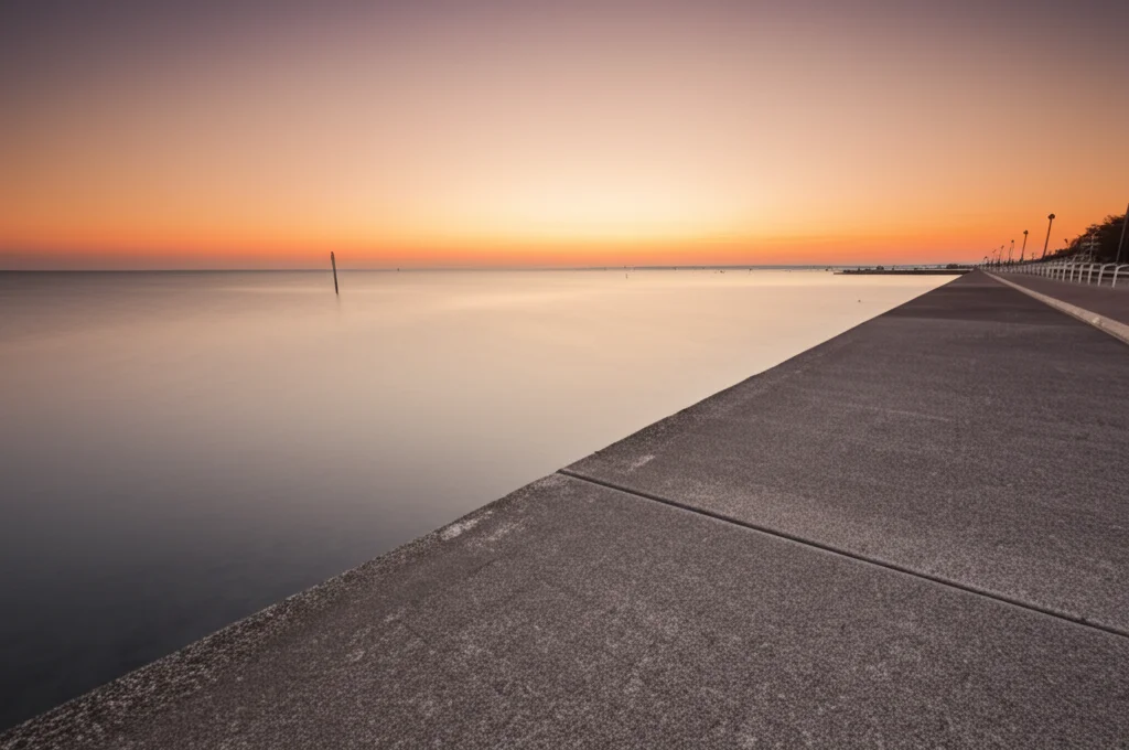 Fotografia grandangolare, 20mm, di un'alta marea primaverile che raggiunge il limite di una passeggiata lungomare in cemento al tramonto. L'acqua è calma ma molto alta, quasi a livello della passeggiata. Il cielo riflette colori caldi sull'acqua. Focus nitido sull'interfaccia acqua-cemento, lunga esposizione per acqua setosa, atmosfera suggestiva.