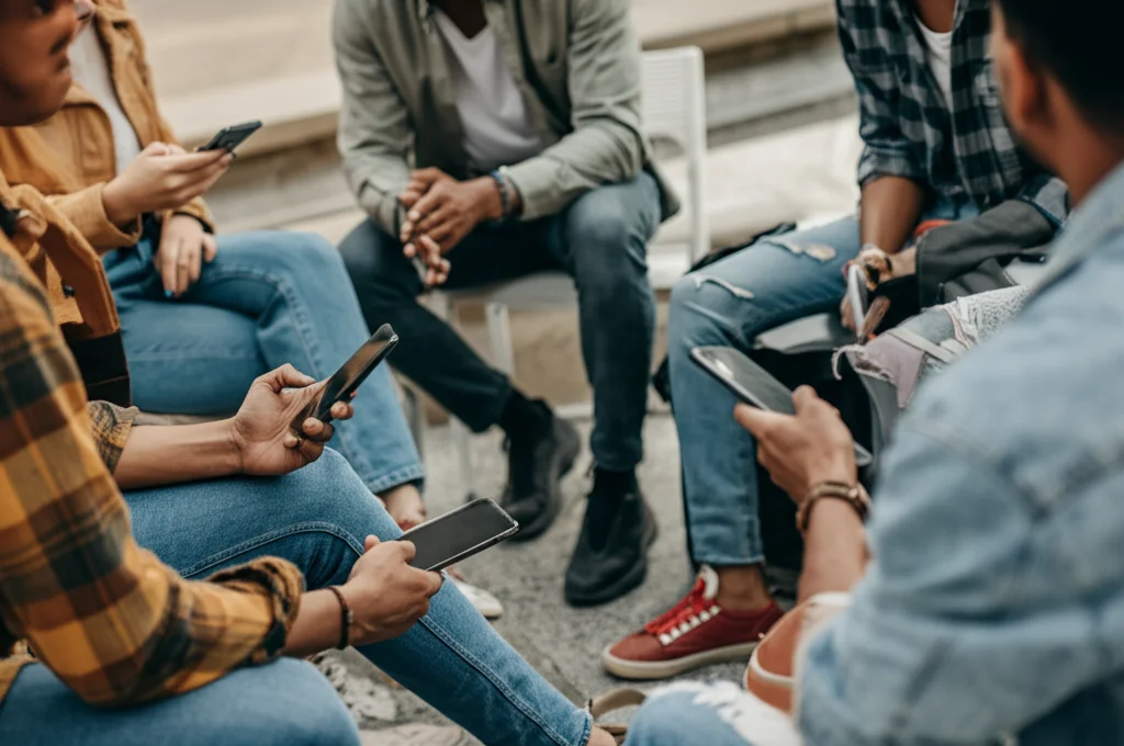 Un gruppo diversificato di giovani adulti (18-24 anni) seduti informalmente in cerchio, forse in un campus universitario o in un parco. Stanno discutendo animatamente ma in modo positivo, alcuni guardano i loro smartphone. L'atmosfera è collaborativa e di supporto. Prime lens, 35mm, depth of field, luce naturale pomeridiana.