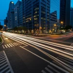 Fotografia aerea di un incrocio stradale trafficato al crepuscolo in una città moderna, le scie luminose delle auto creano un senso di movimento e potenziale pericolo. Obiettivo grandangolare 24mm, lunga esposizione per scie luminose, focus nitido sull'incrocio.