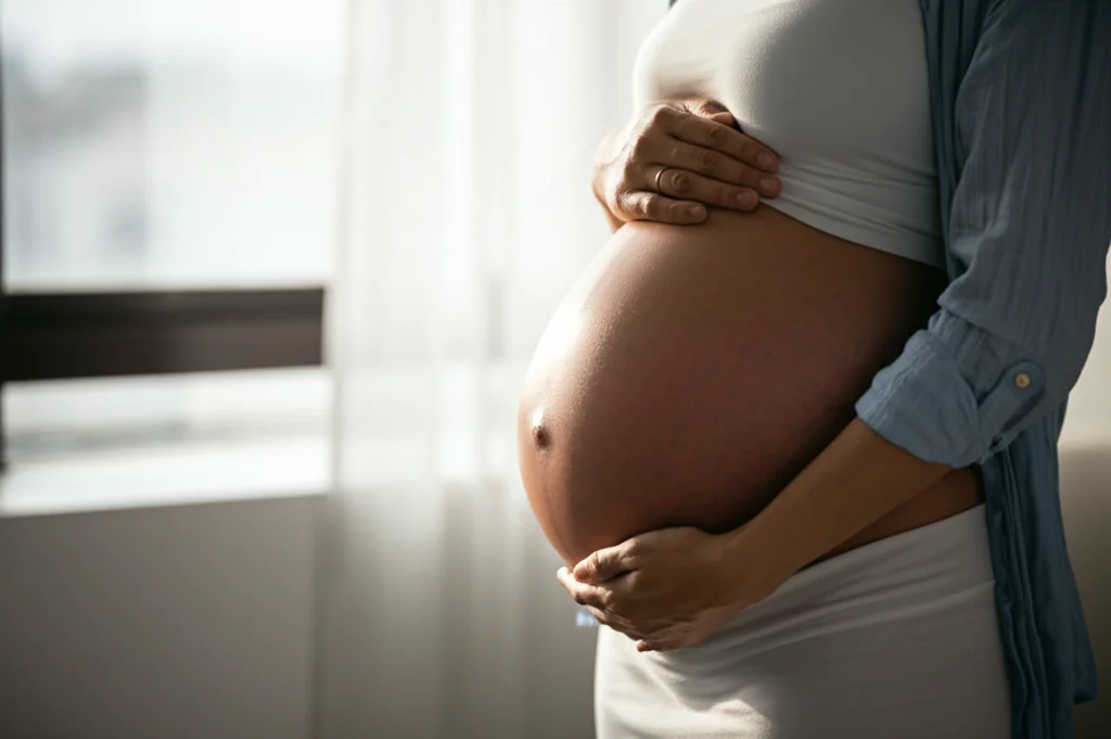 Primo piano fotografico di una donna incinta che sorride con speranza, tenendo delicatamente il pancione. Luce calda e naturale proveniente da una finestra, obiettivo 50mm, profondità di campo ridotta per isolare il soggetto e creare uno sfondo morbido, simboleggia un futuro più sano e la positività dell'intervento.