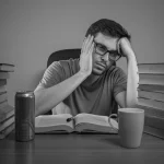 Ritratto di uno studente di medicina stanco seduto a una scrivania piena di libri, con una tazza di caffè e una lattina di energy drink visibili, obiettivo 35mm, profondità di campo, illuminazione soffusa da studio, bianco e nero.