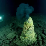 Suggestiva immagine subacquea del fondale del vulcano Kolumbo, con un camino idrotermale ('black smoker') attivo che emette un denso pennacchio nero di fluidi ricchi di minerali. Le luci potenti di un veicolo sottomarino (ROV) illuminano la scena oscura, rivelando le aspre rocce vulcaniche circostanti e la complessa struttura mineralizzata del camino. Obiettivo grandangolare 12mm, lunga esposizione per catturare il movimento etereo del fumo nero, messa a fuoco nitida sulla struttura del camino e sulle rocce circostanti.