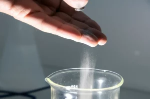 Immagine concettuale che mostra una mano umana che lascia cadere una polvere grigio-metallica scintillante (simbolo delle Terre Rare) in acqua limpida contenuta in un becher da laboratorio, l'acqua inizia a intorbidirsi leggermente nella zona di caduta. Illuminazione da laboratorio, obiettivo 50mm, profondità di campo media per mantenere a fuoco sia la mano che l'acqua.